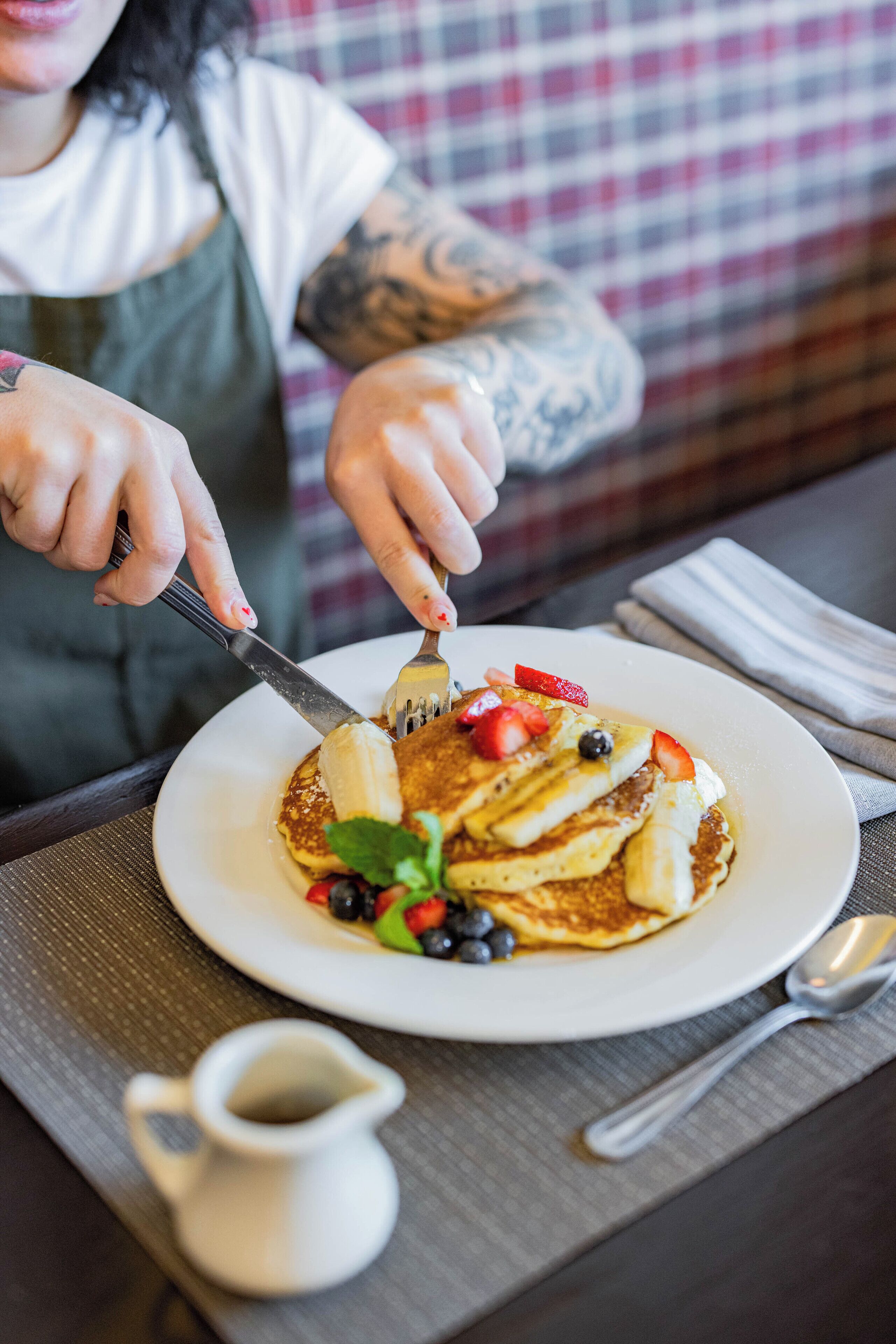 A person cutting a pancake topped with strawberries, blueberries, and banana slices, with a small pitcher of syrup on the table.