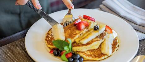 A person cutting a pancake topped with strawberries, blueberries, and banana slices, with a small pitcher of syrup on the table.
