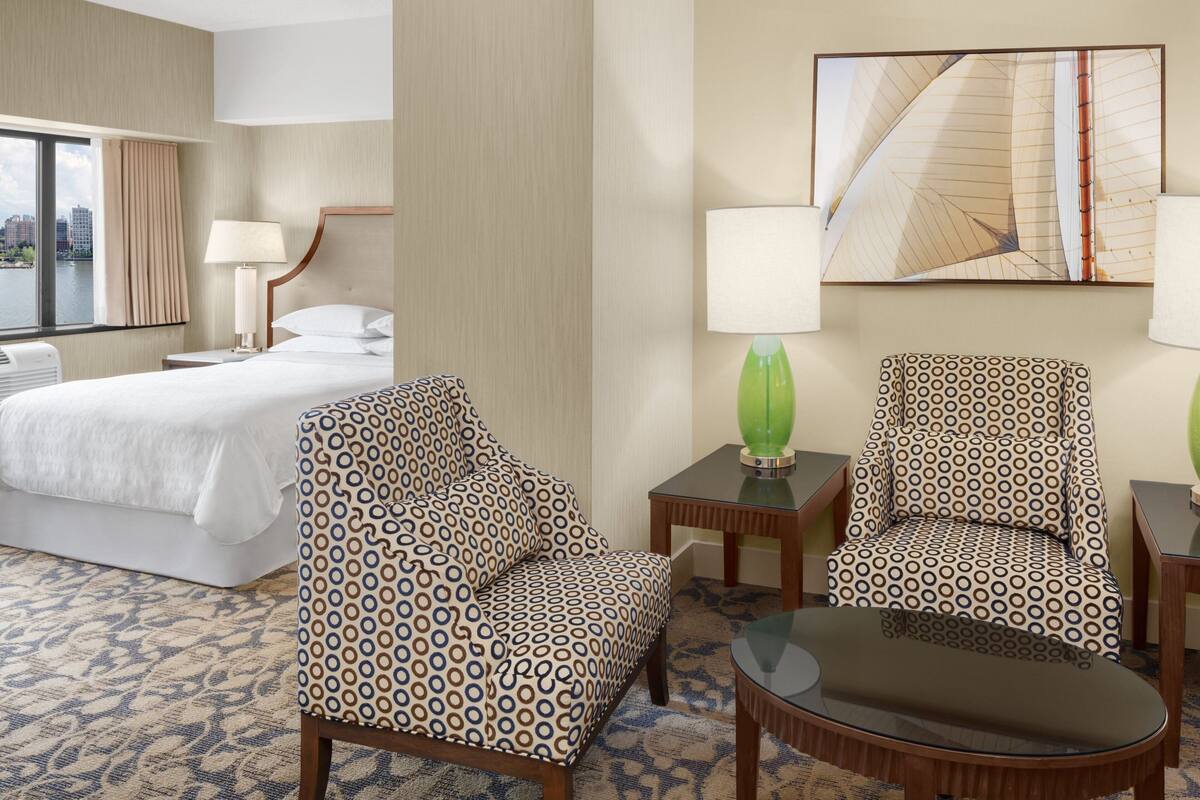 A hotel room with a bed, two patterned armchairs, a small table, and a painting on the wall.