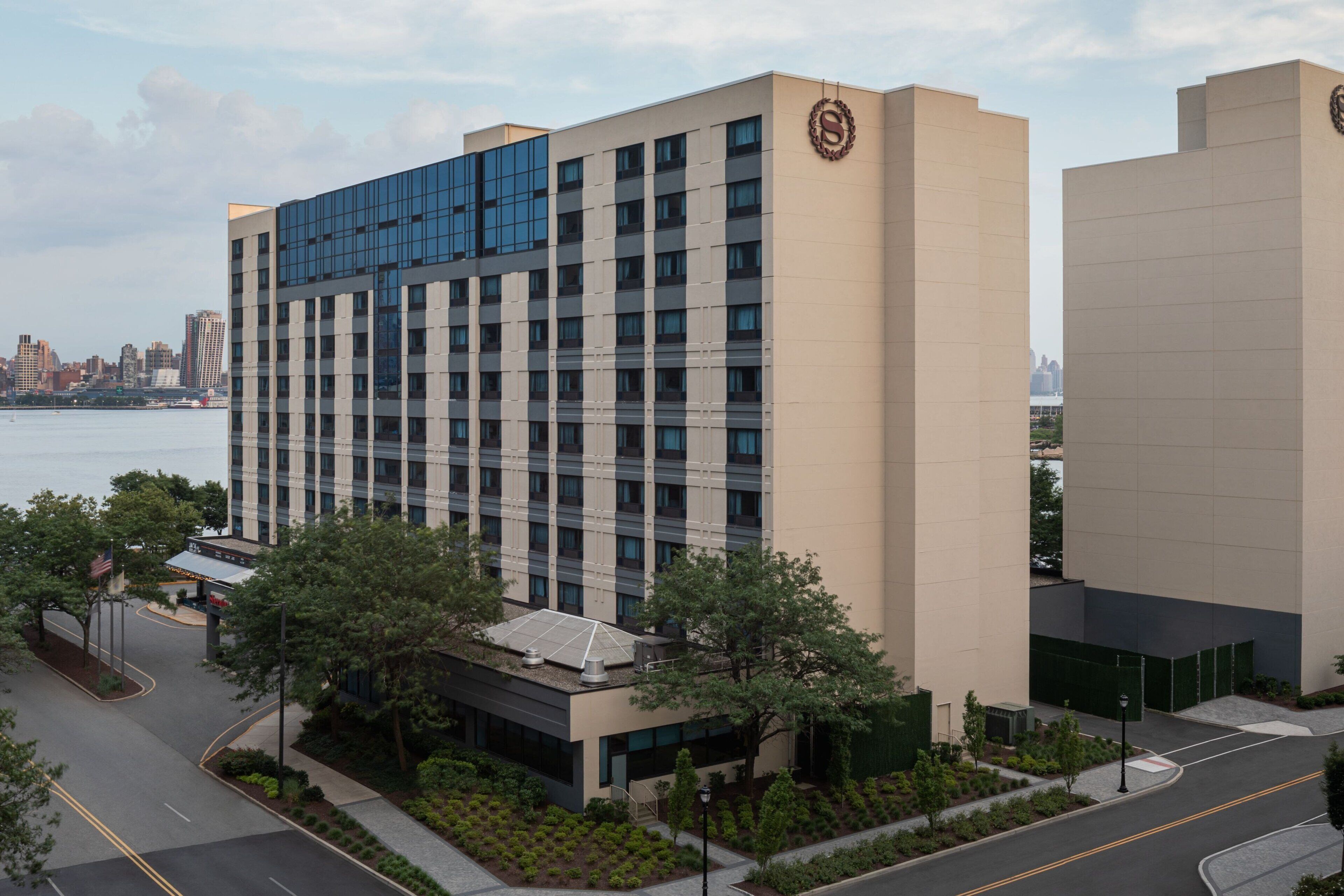 A modern hotel with a glass facade, a parking area, and a view of the city skyline.