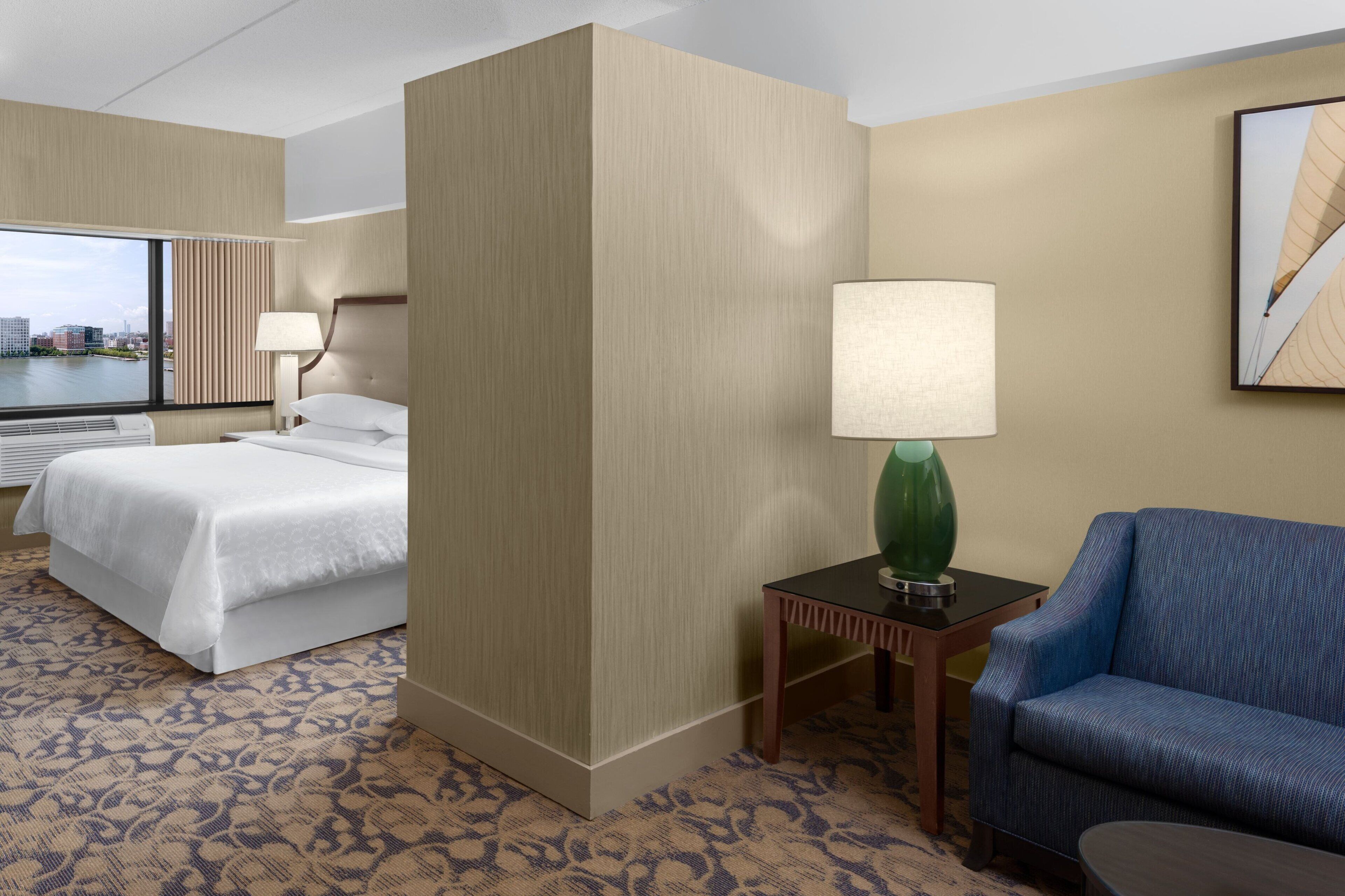 A hotel room with a bed, a desk, a chair, a lamp, and a city view through the window.