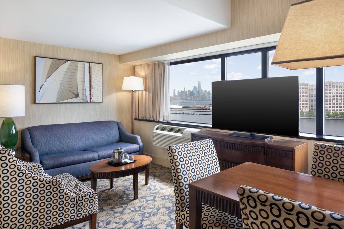 A hotel room with a large window, a sofa, a dining table, patterned chairs, a flat-screen TV, and a decorative mirror on the wall.