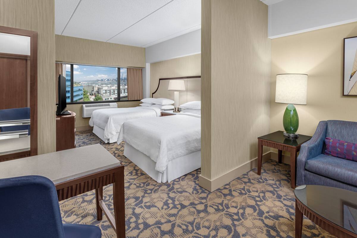 A hotel room with two beds, a desk, a chair, a TV, and a view of the city.
