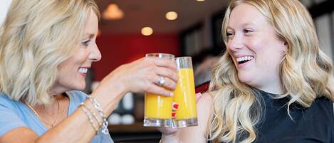 Zwei Personen stoßen mit Orangensaft an einem Tisch an, auf dem ein Gebäck und eine Tasse stehen.