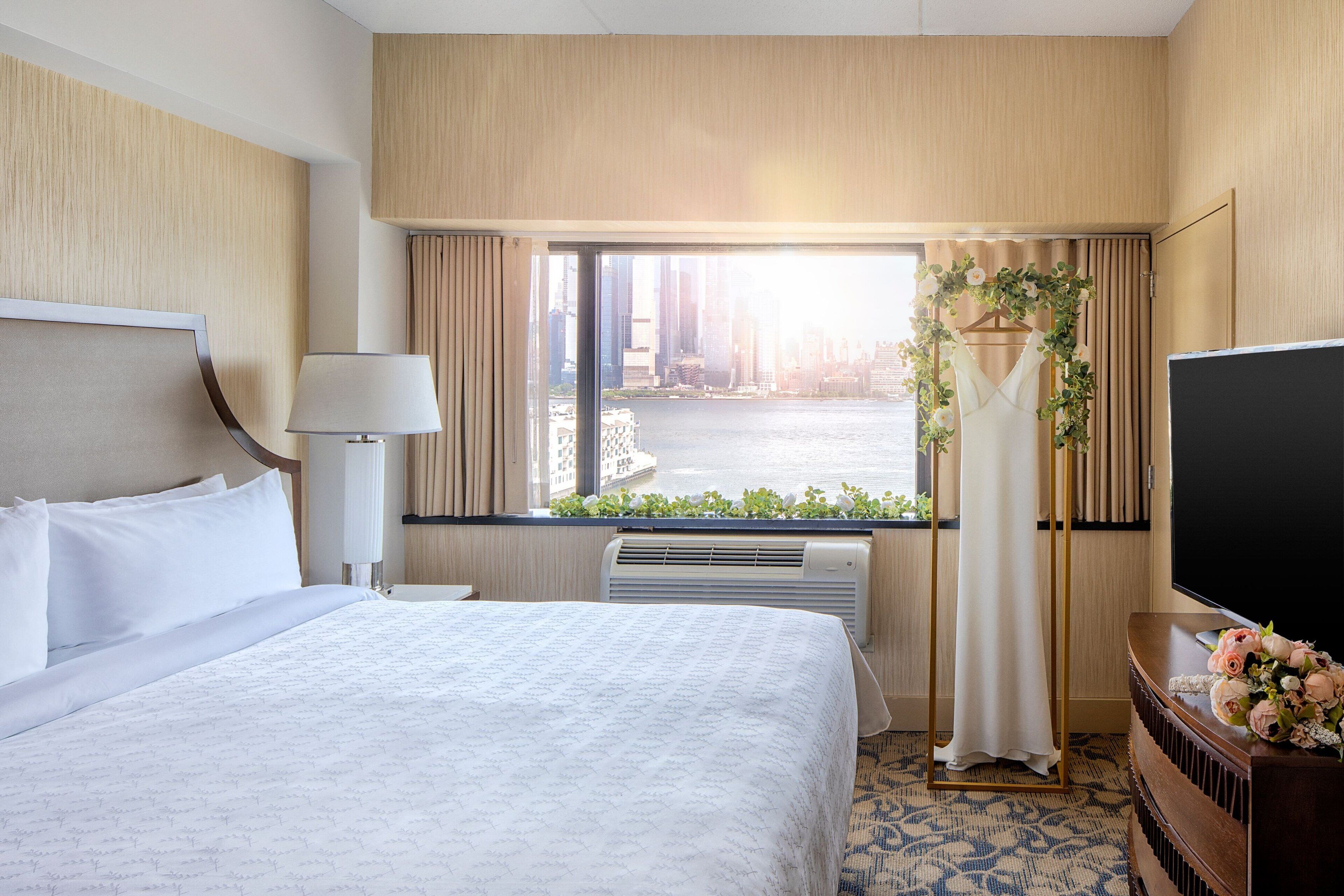 A hotel room with a large bed, a television, a window with curtains, and a view of a cityscape.