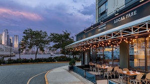 Un área de comedor al aire libre con mesas y sillas, y vista al horizonte de la ciudad. Un letrero dice 'LOCAL FARE'.