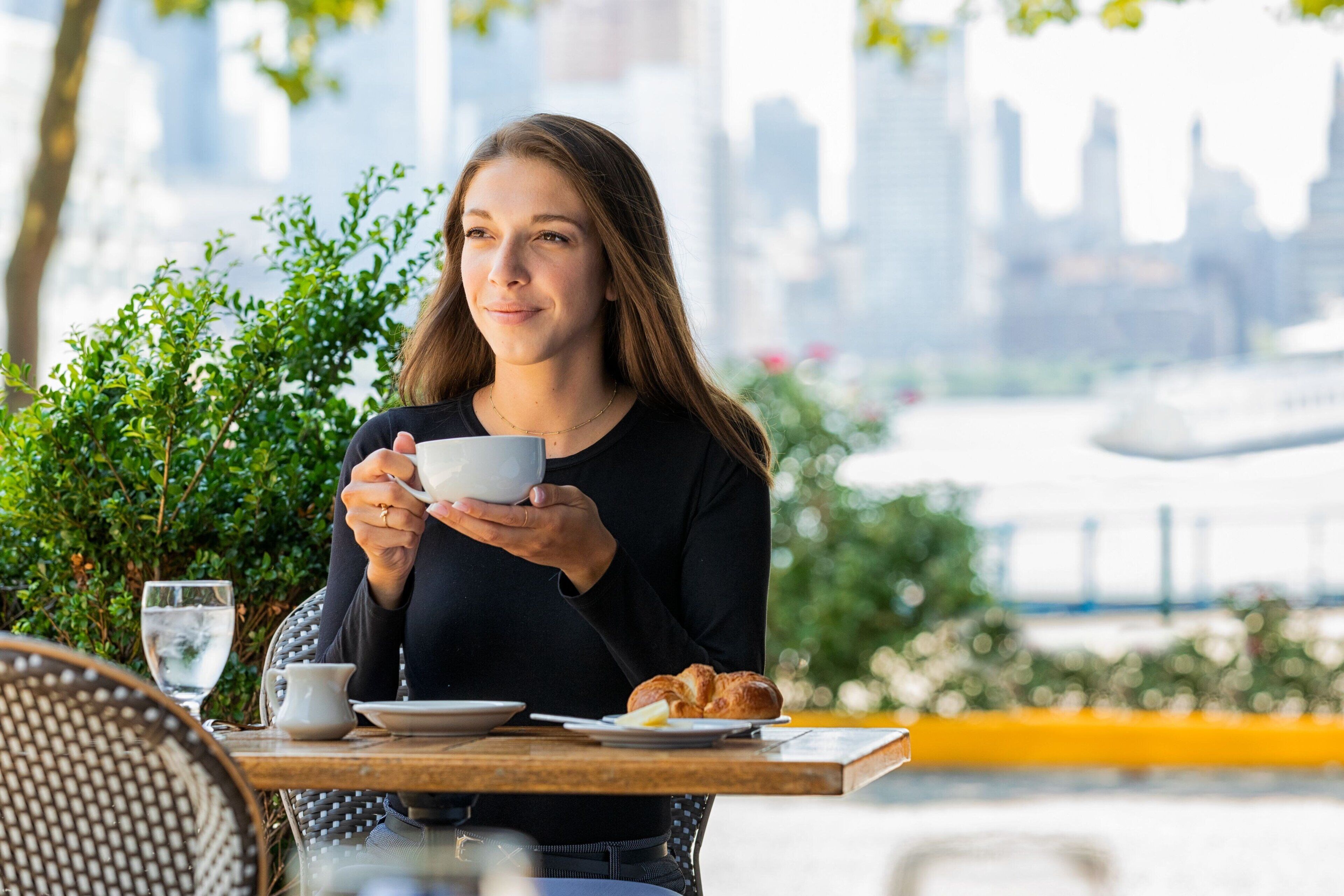 A person holding a cup at an outdoor table with a cityscape in the background.