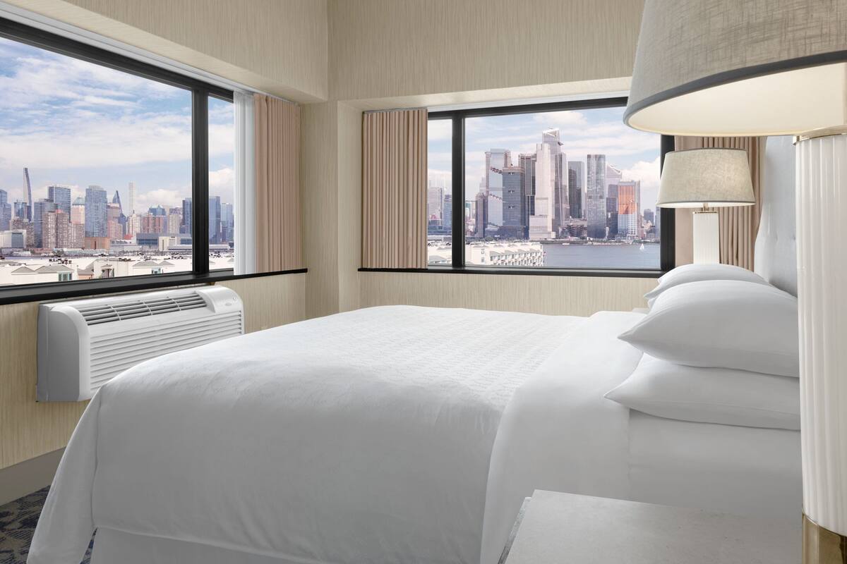 A hotel room with a large bed, a view of a city skyline, and a window with curtains.