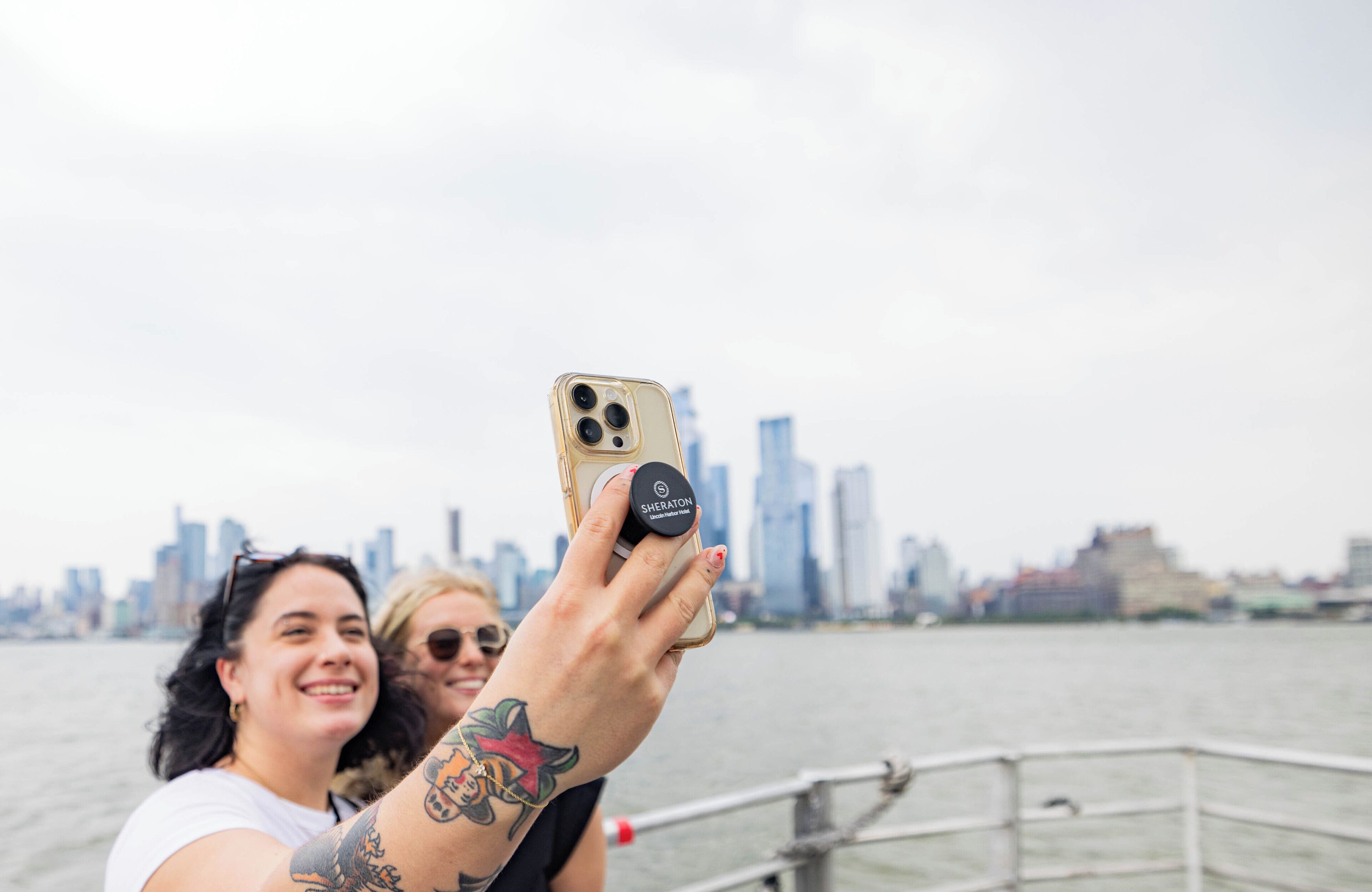 Una persona sosteniendo un teléfono inteligente con una funda Sheraton, tomándose un selfie con un horizonte urbano de fondo.