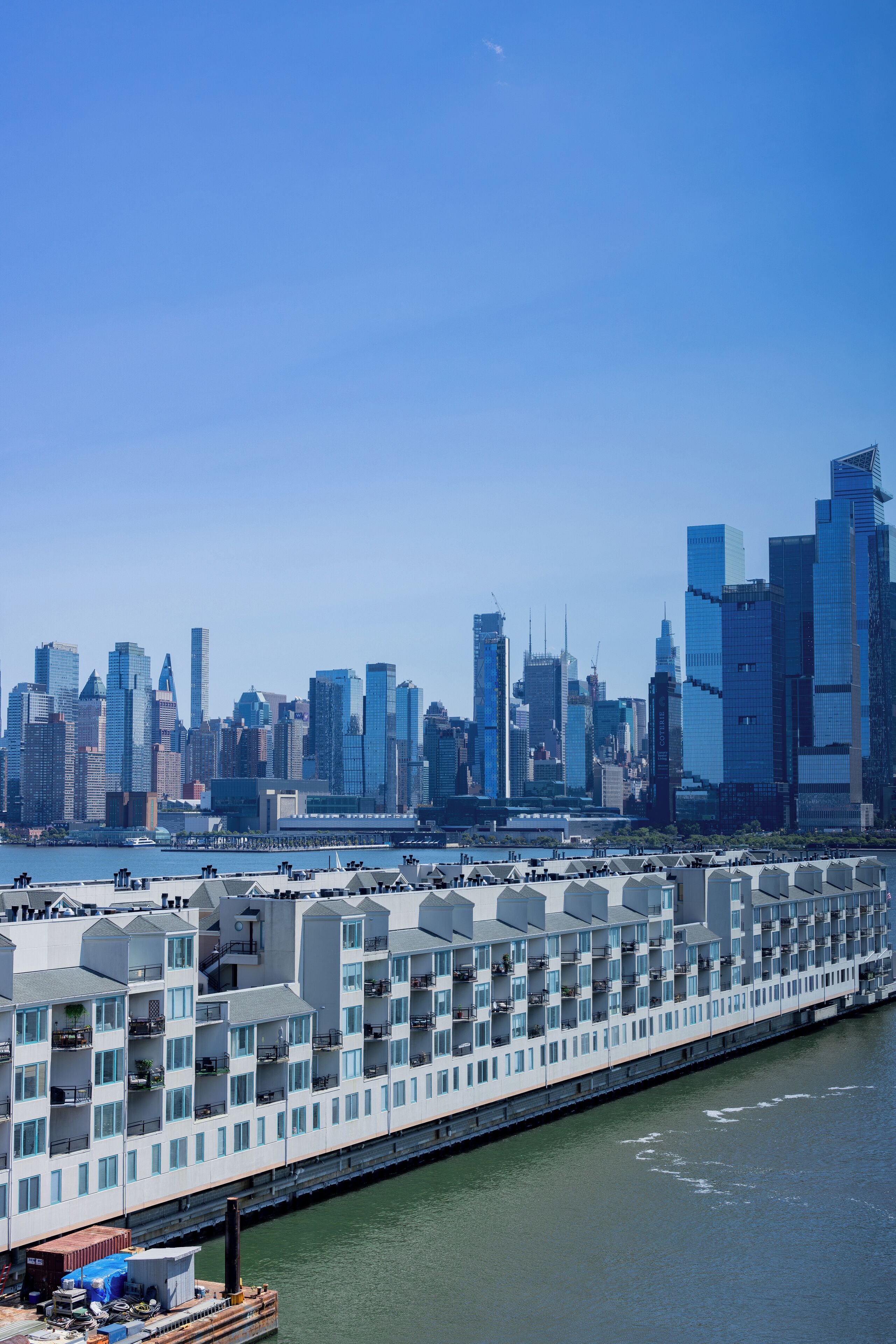 A waterfront hotel with multiple balconies, a river view, and a city skyline in the background.