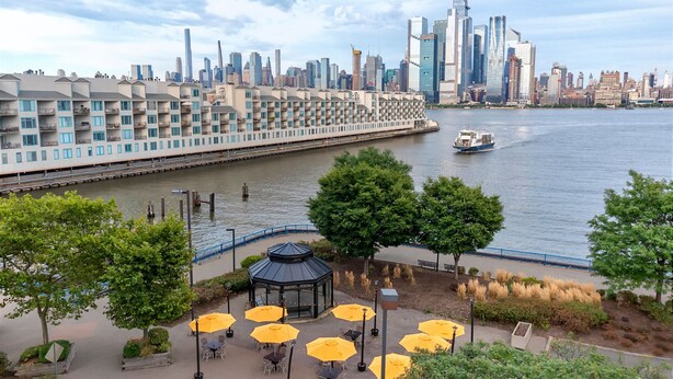 Un patio junto al río con mesas al aire libre y un pabellón, con vistas a un horizonte urbano de edificios modernos y un barco en el agua.