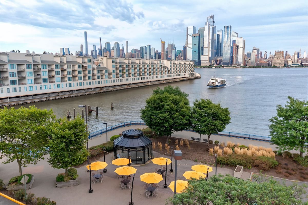 A riverside patio with outdoor seating and a gazebo, overlooking a city skyline with modern buildings and a boat on the water.