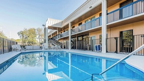 Una piscina con superficie de agua azul cristalina, rodeada por una terraza blanca. Hay sillas de descanso y una escalera que lleva a un edificio de varios niveles con balcones.