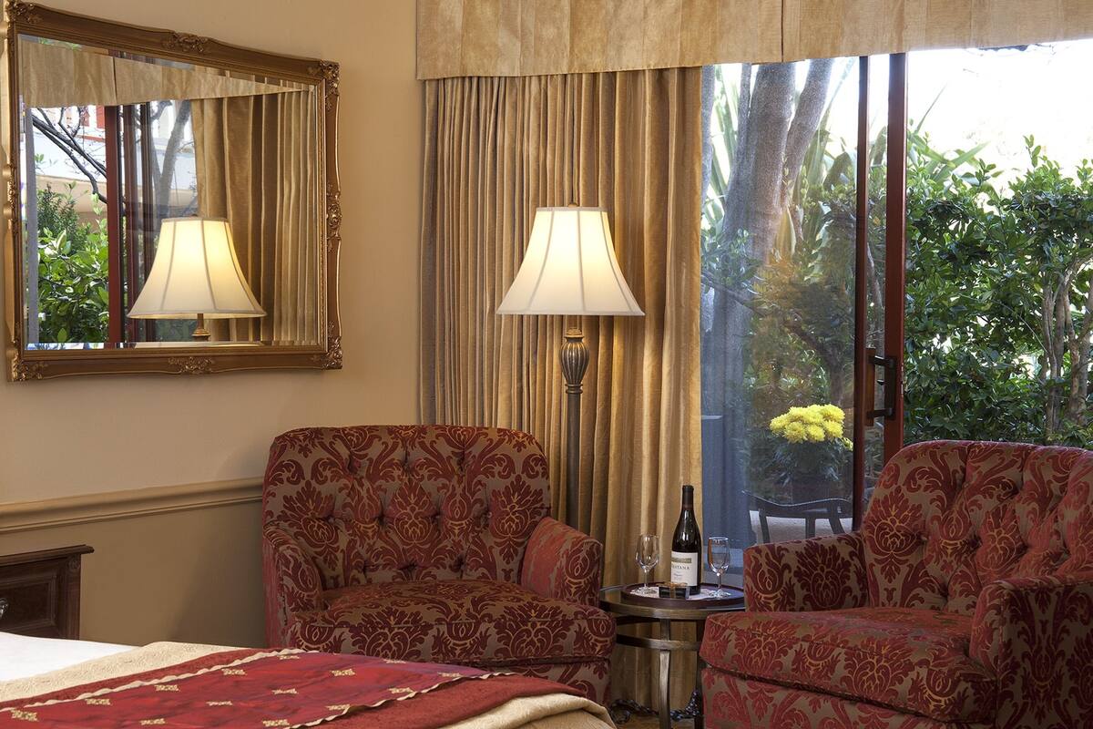 A hotel room with a bed, two patterned armchairs, a table with a bottle and glasses, a mirror, and a view of greenery through large windows.