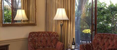 Habitación de hotel con una cama, dos sillones estampados, una mesa con una botella y copas, un espejo y vistas a zonas verdes a través de grandes ventanales.