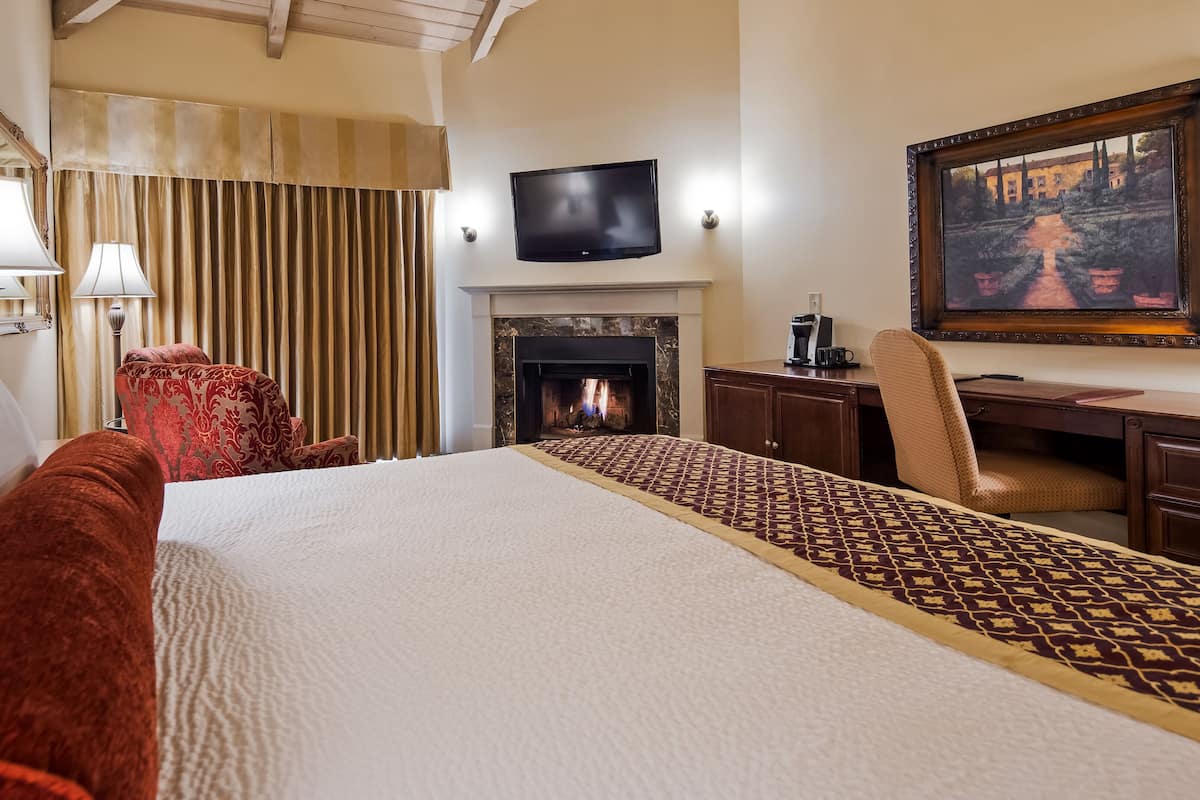 A hotel room with a large bed, a desk, a chair, a fireplace, and a television on the wall.