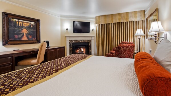 A hotel room with a bed, a desk, a chair, a fireplace, a television, and a window with curtains. - Victorian Inn (Monterey)