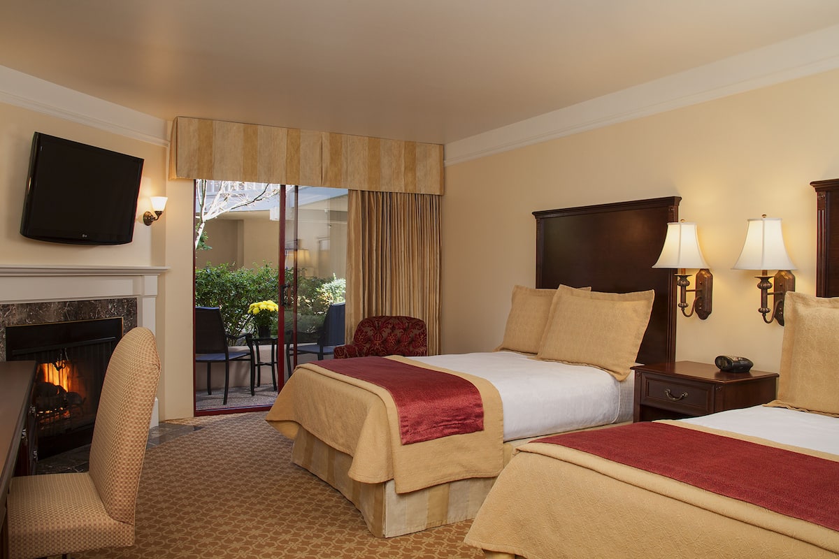 A hotel room with a fireplace, two beds with red and beige bedding, a TV mounted on the wall, and a view of the outdoors.