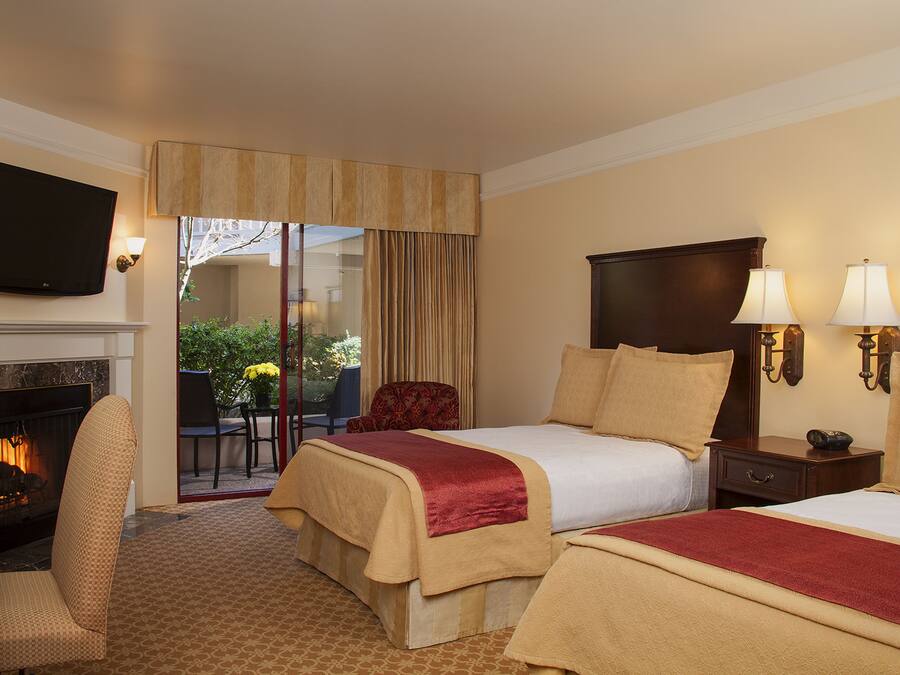 A hotel room with a fireplace, two beds with red and beige bedding, a TV mounted on the wall, and a view of the outdoors.