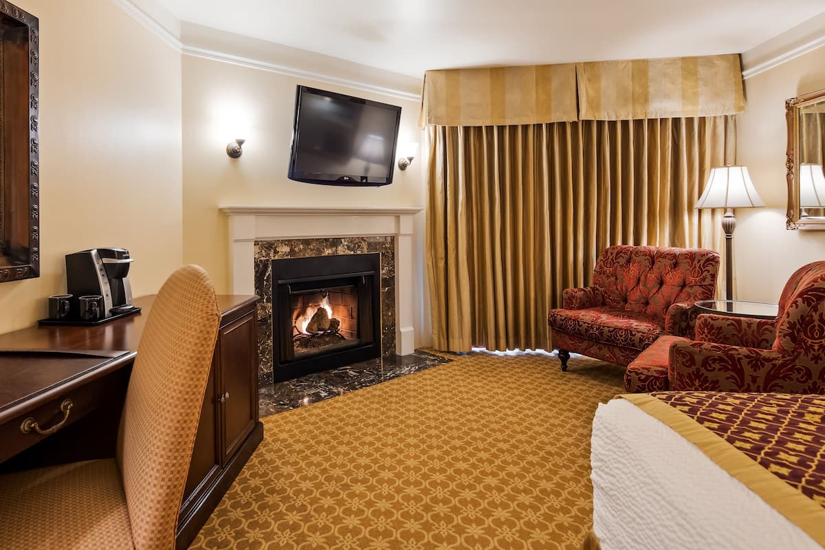 A hotel room with a fireplace, a television, a desk with a coffee maker, and a comfortable seating area with a patterned carpet.
