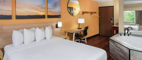 A hotel room with a large bed, a desk, a chair, a bathroom with a bathtub, and two sunset paintings on the wall.