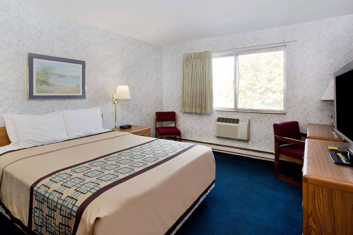 A hotel room with a bed, a desk, a chair, a TV, a window with curtains, and a painting on the wall.