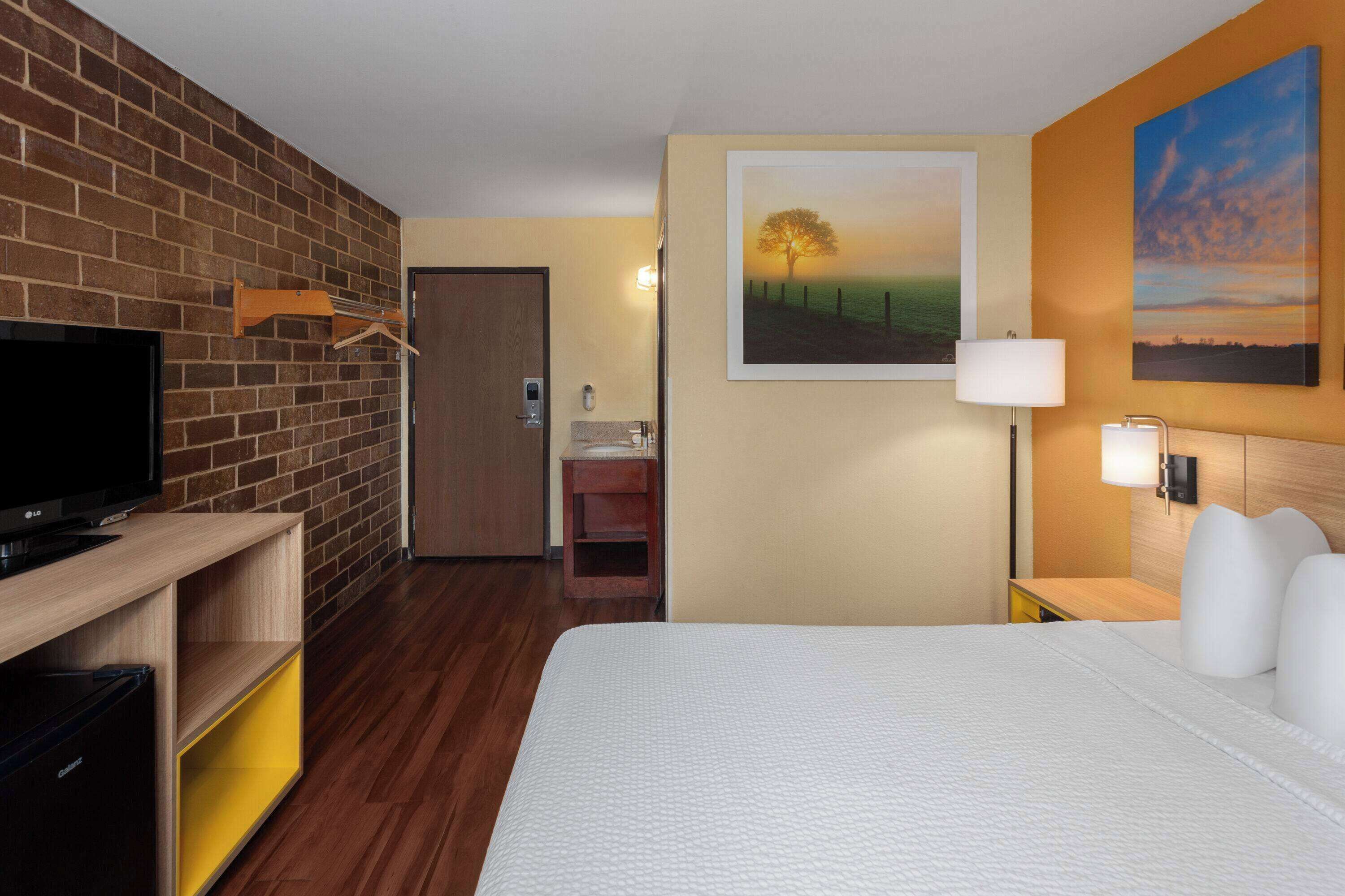 A hotel room with a brick wall, a flat-screen TV, a bed with white bedding, a painting of a tree, and a lamp.