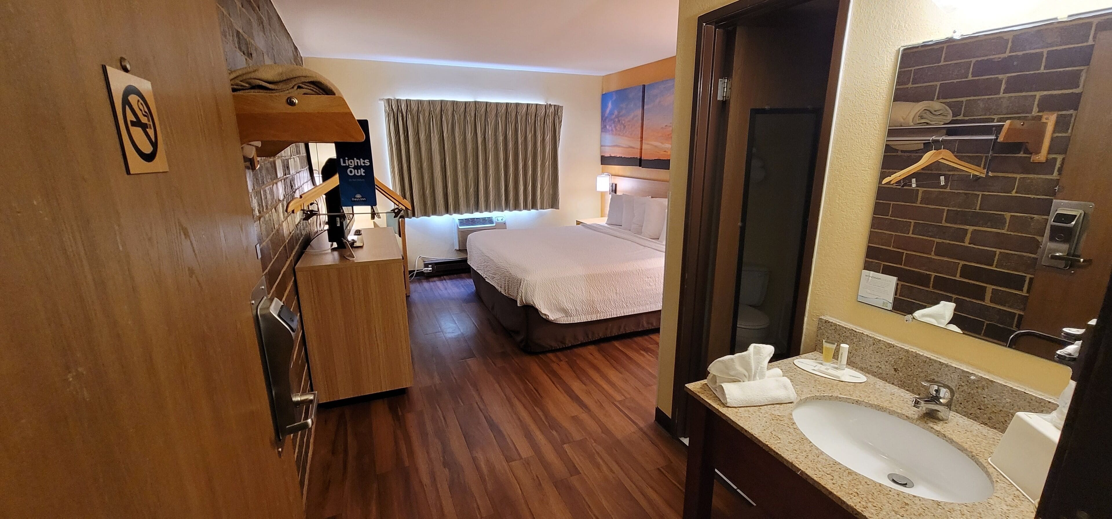 A hotel room with a wooden floor, a bed with white bedding, a bathroom with a sink and mirror, and a window with curtains.