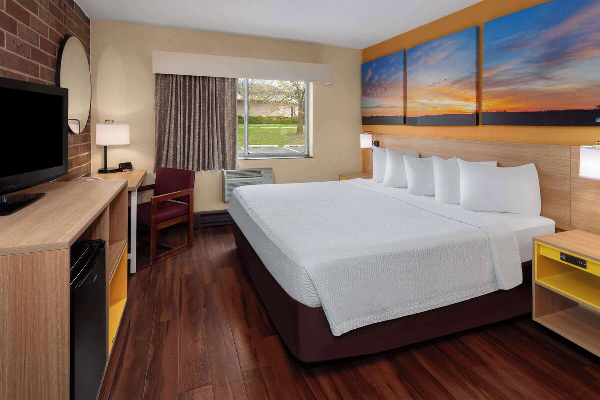 A hotel room with a large bed, a desk with a television, a chair, a window with curtains, and a painting on the wall.