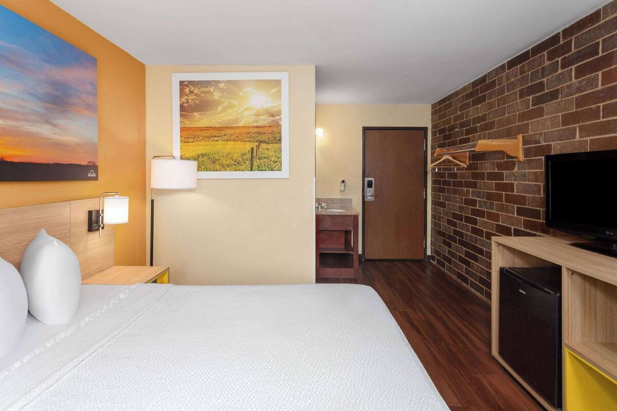 A hotel room with a bed, a TV, a brick wall, and a painting of a sunset.