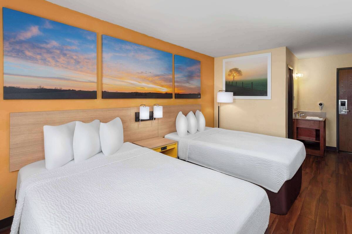 A hotel room with two beds, a wooden headboard, a nightstand with a lamp, a small table with a mirror, and artwork on the wall.