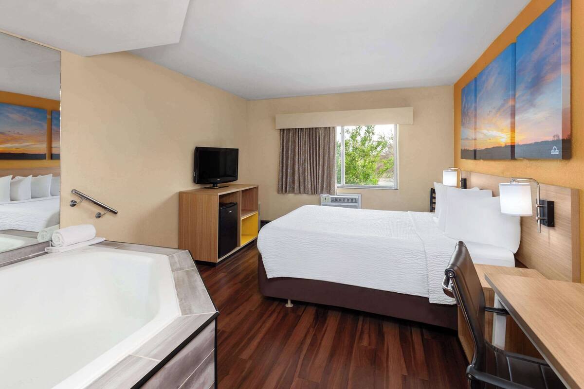 A hotel room with a large bed, a desk, a TV, a bathtub, and a window with curtains.