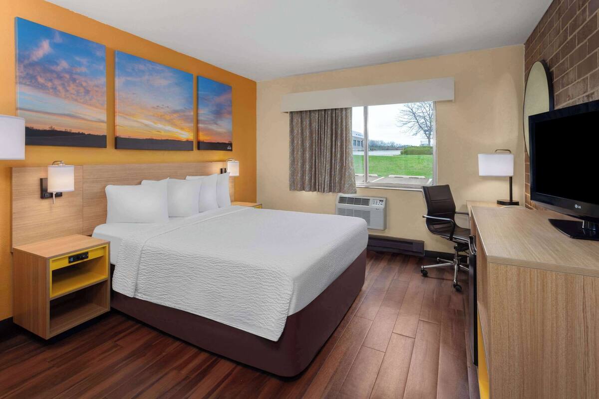 A hotel room with a large bed, a desk with a TV, a window with curtains, and artwork on the wall.