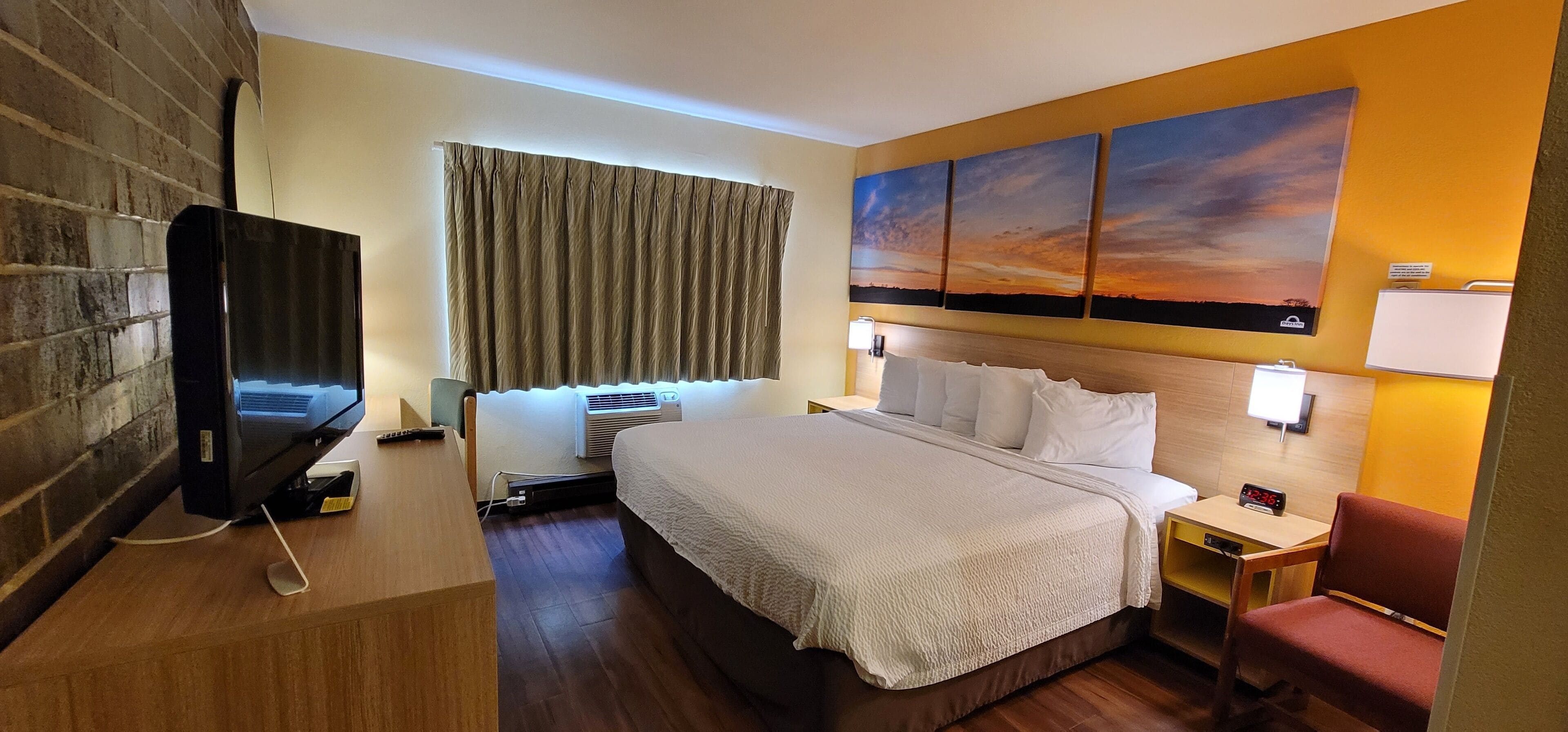 A hotel room with a bed, a TV, a wall-mounted heater, a window with curtains, and a sunset painting on the wall.