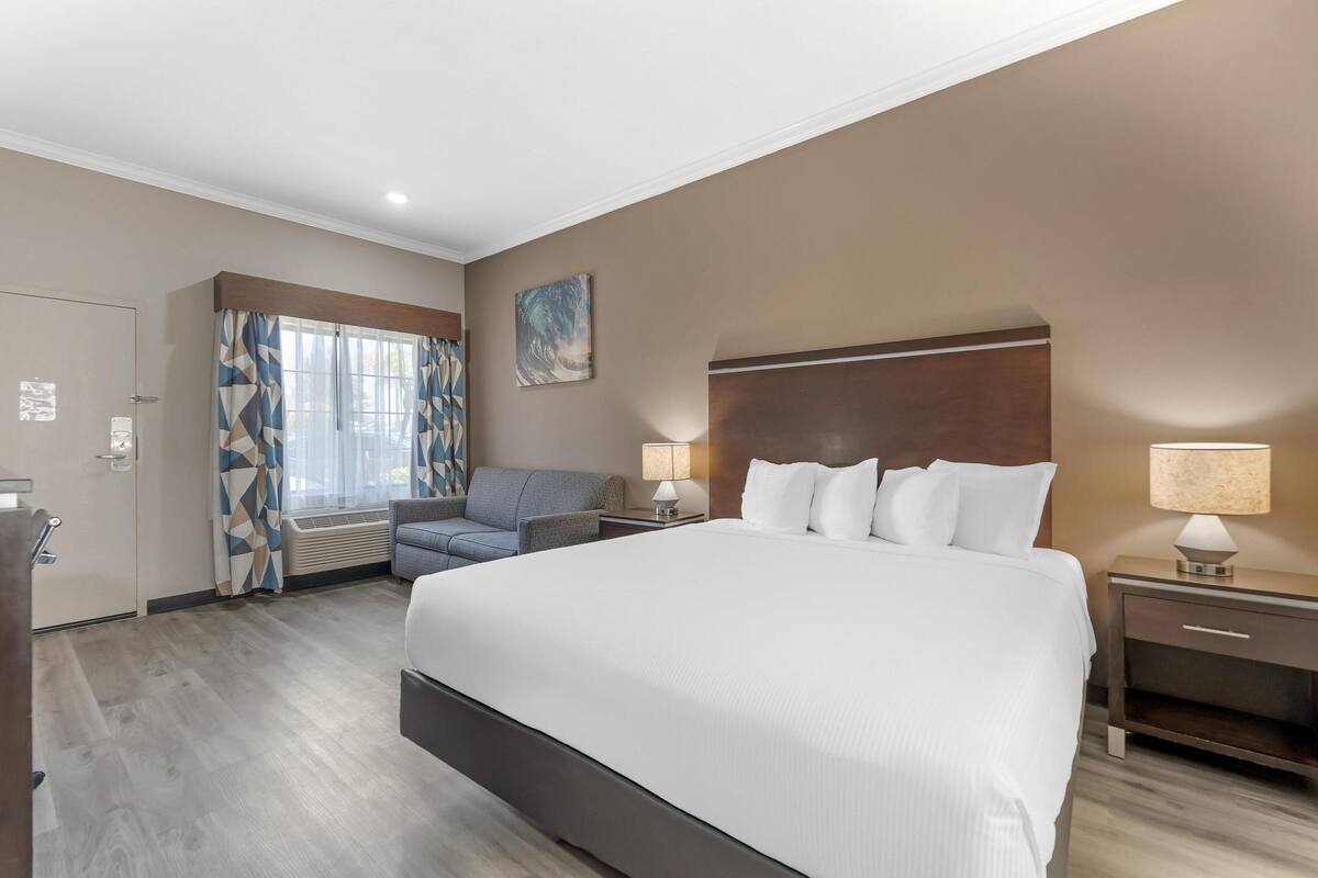 Une chambre d’hôtel avec un grand lit, un bureau, une chaise, une fenêtre avec des rideaux et un tableau accroché au mur.