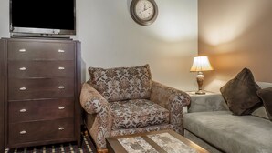 A living room with a television on a cabinet, a patterned armchair, a sofa, and a lamp on a side table. - Rodeway Inn Allentown South (Allentown)