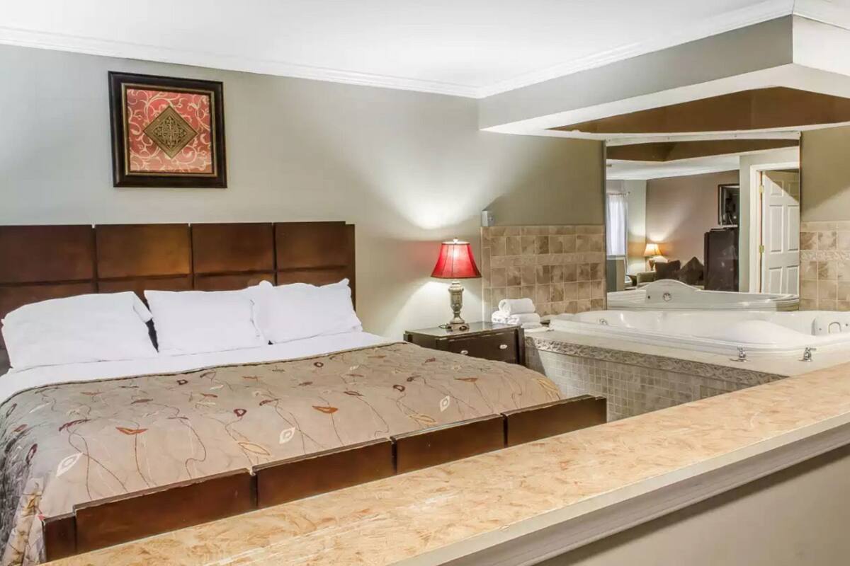 A hotel room with a bed, a nightstand, a red lamp, a large mirror, and a bathtub.