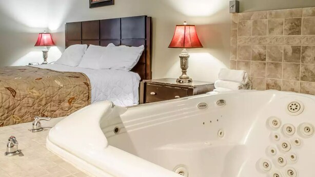 A hotel room with a large white bathtub, a double bed with a brown headboard, two bedside lamps, and a tiled wall. - Rodeway Inn Allentown South (Allentown)