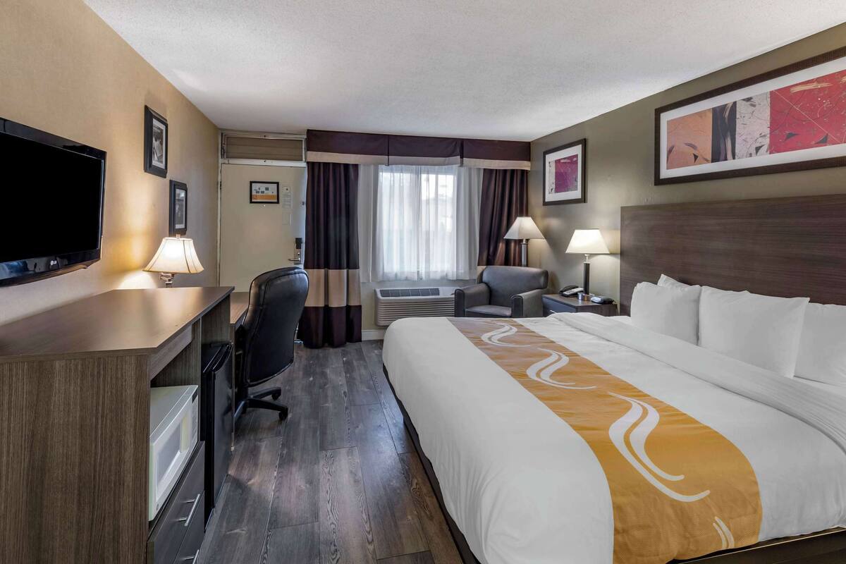 A hotel room with a large bed, a desk with a flat-screen TV, a chair, a desk lamp, and a window with curtains.
