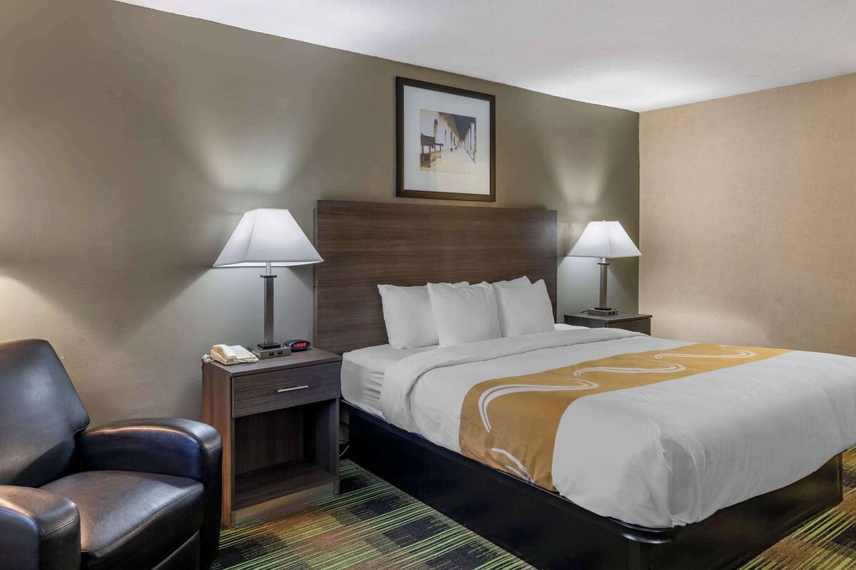 A hotel room with a bed, two bedside lamps, a chair, a nightstand, a framed picture on the wall, and a carpet.