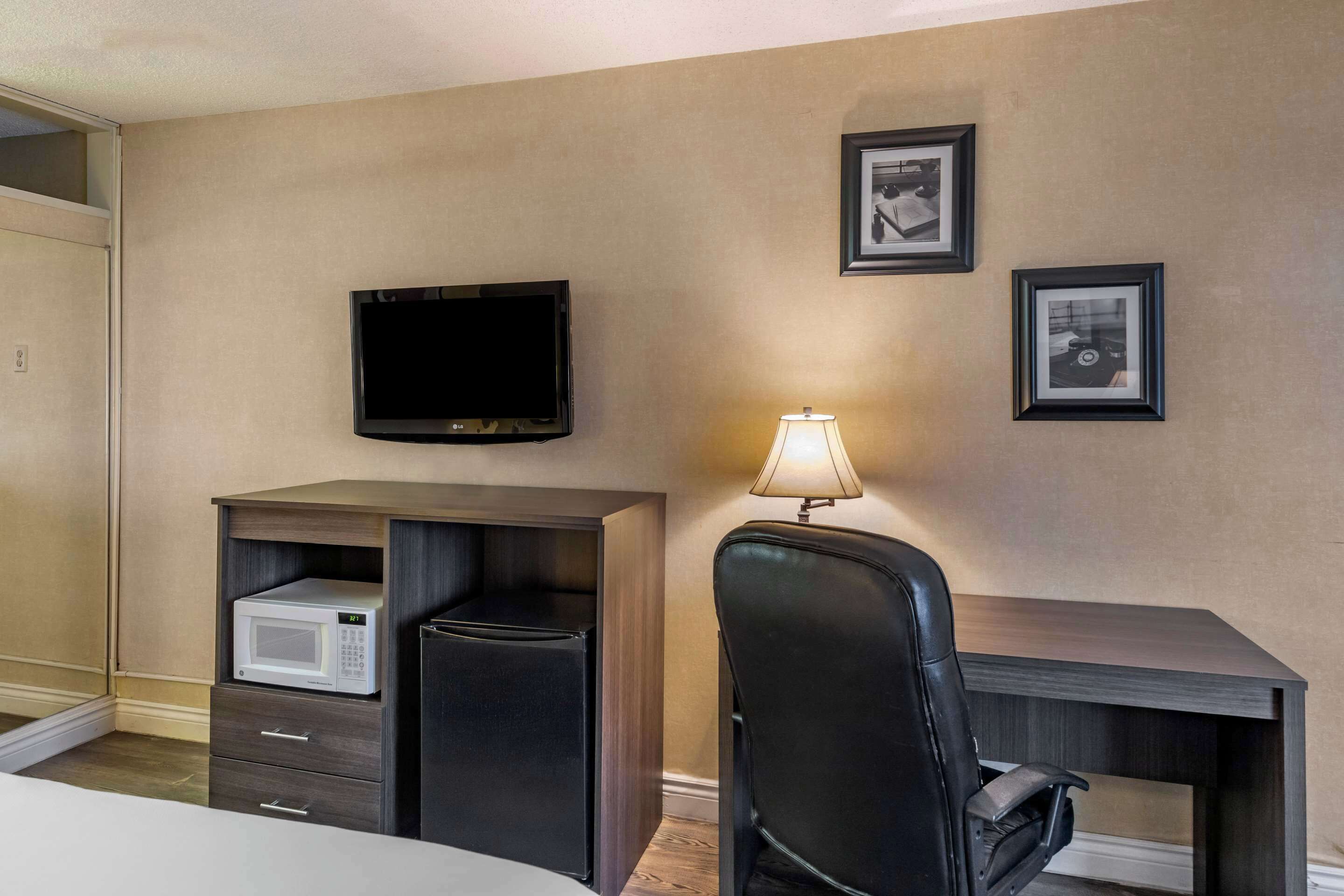 A hotel room with a flat-screen TV mounted on the wall, a desk with a lamp, a microwave, and a chair.