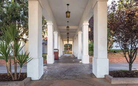 A covered walkway with white columns and hanging lanterns, leading to a fountain.