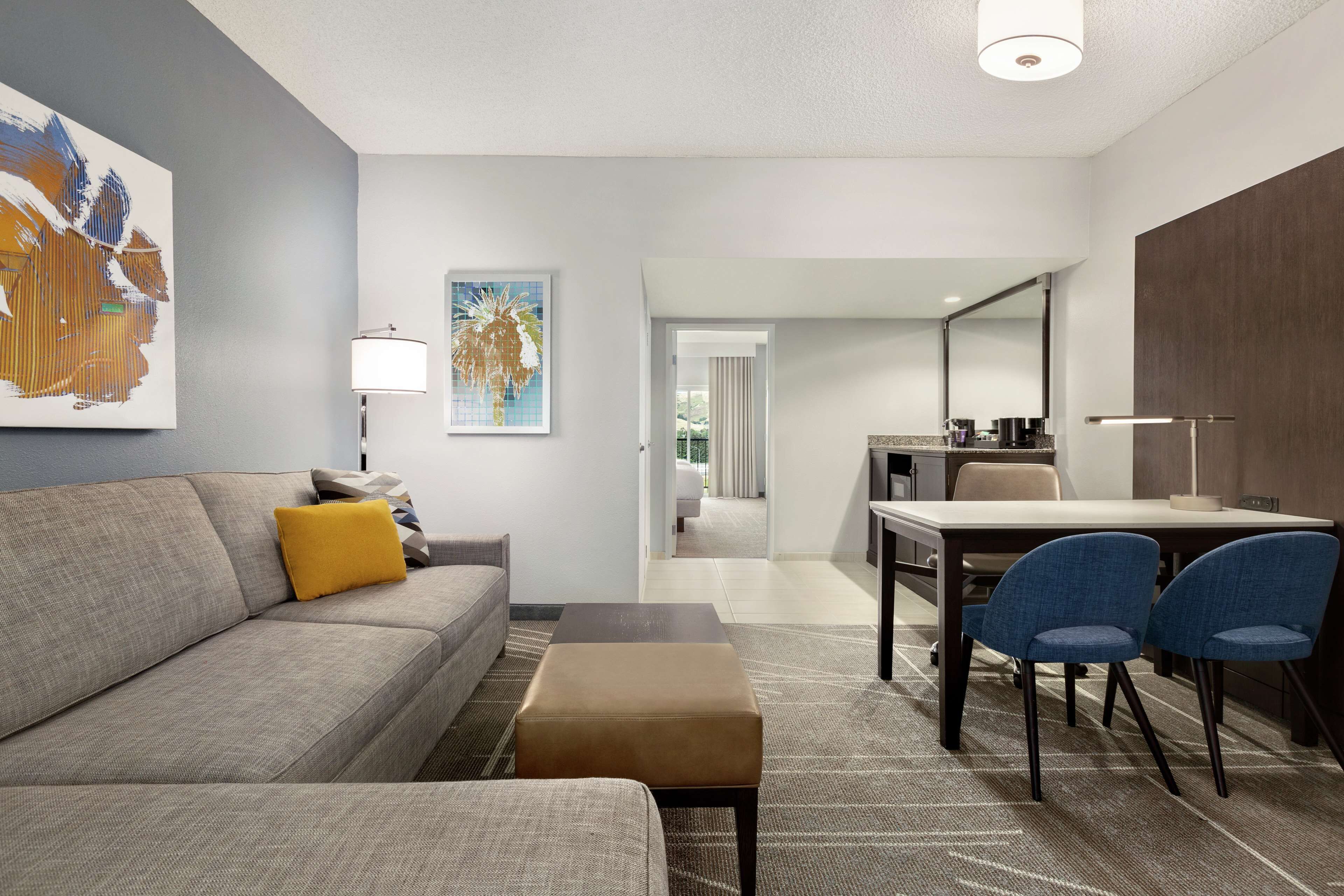 Una habitación de hotel moderna con un sofá gris, un otoman marrón, una mesa de comedor con dos sillas azules y un cuadro grande en la pared.