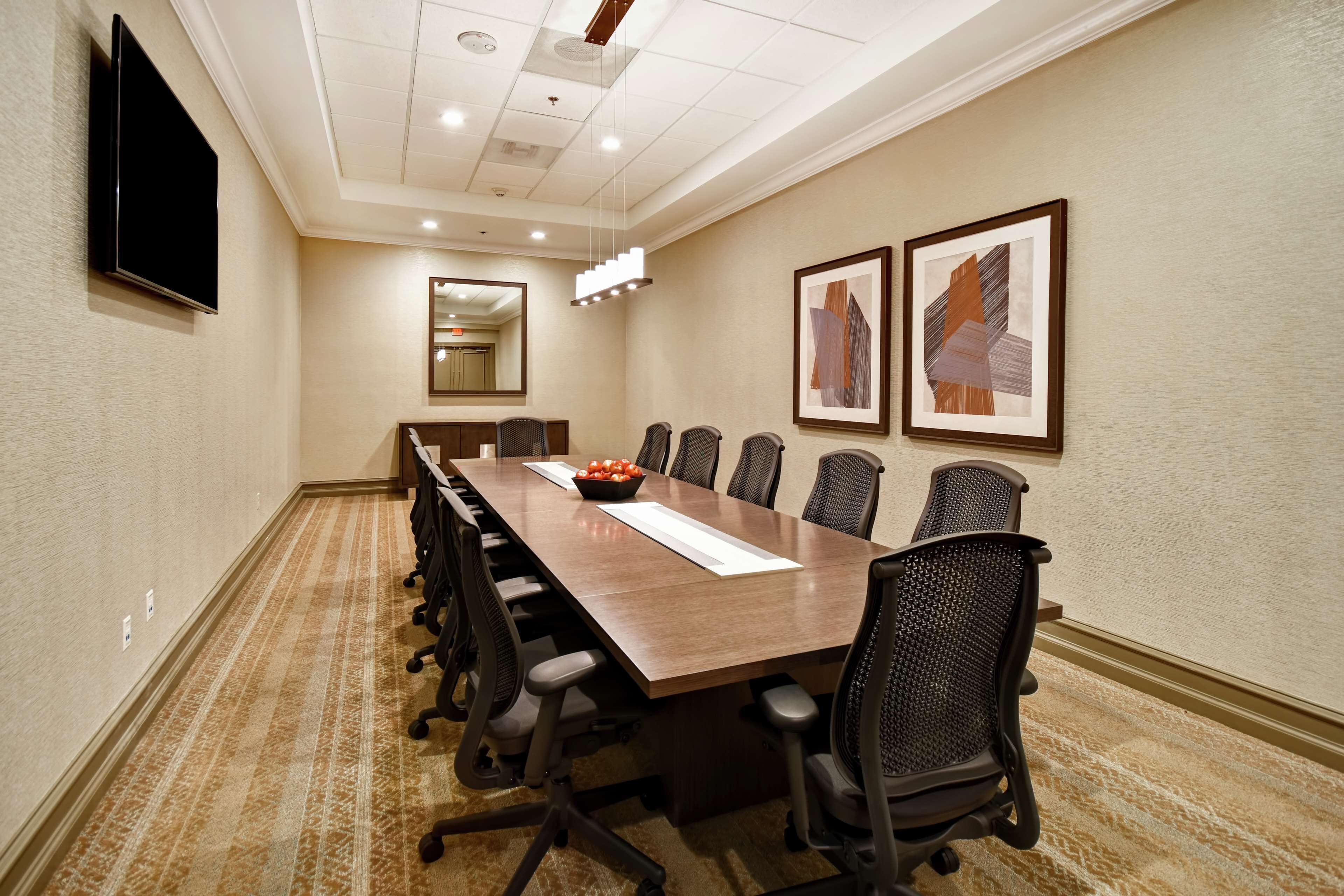 A conference room with a long table, chairs, a flat-screen TV, framed artwork, and a carpeted floor.