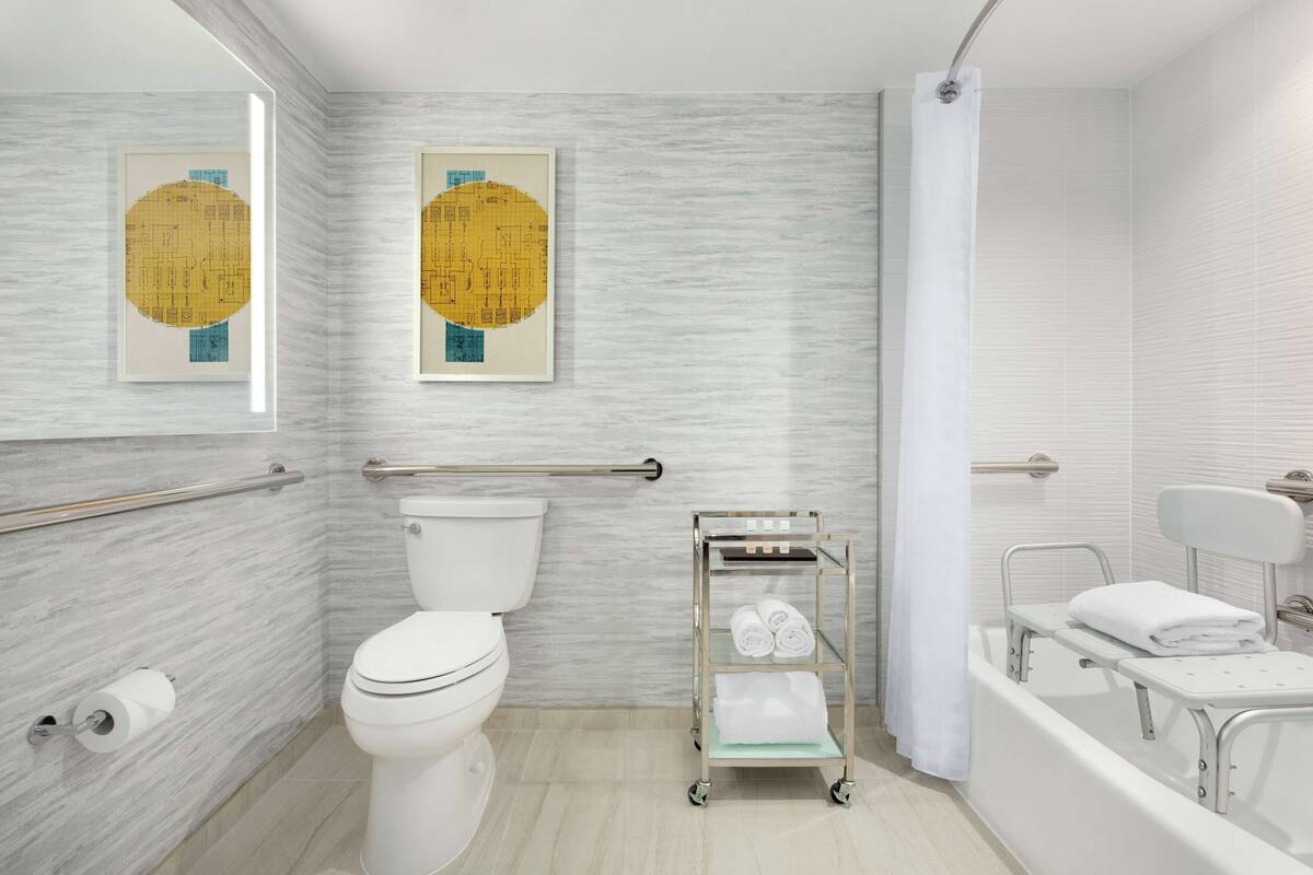 A modern bathroom with a toilet, bidet, and bathtub. There are two framed artworks on the wall, a towel rack, and a cart with towels.
