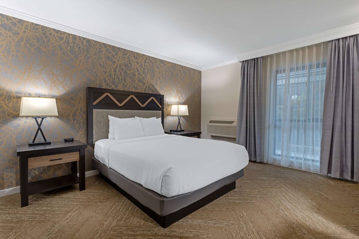 A hotel room with a large bed, two bedside tables with lamps, a headboard with a zigzag design, a wall with a gold pattern, and a window with curtains.