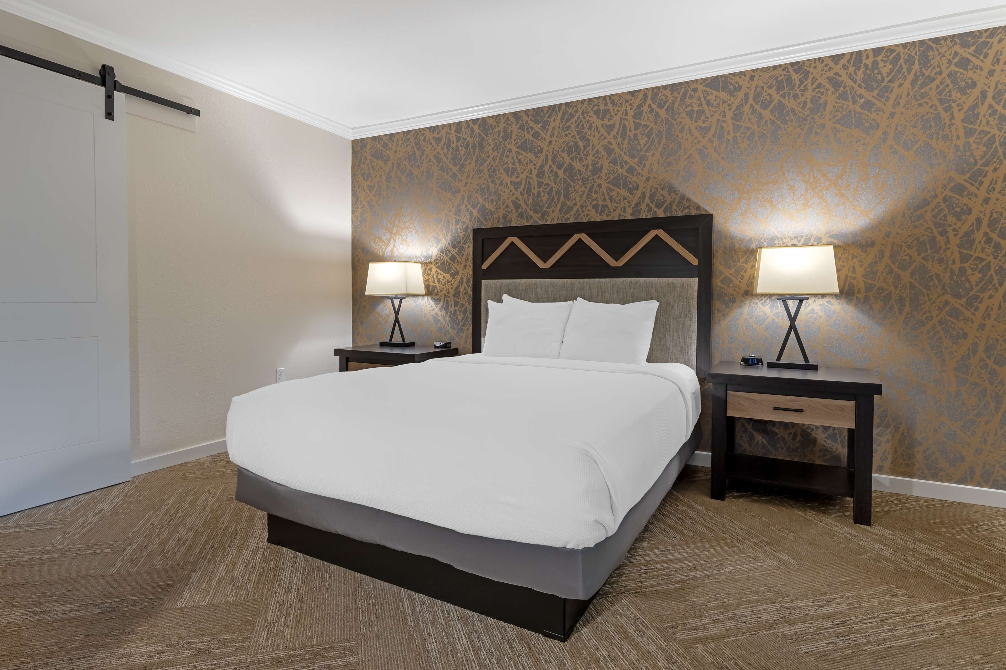 Une chambre d’hôtel avec un grand lit, deux tables de chevet avec des lampes, une tête de lit en bois et un papier peint à motifs.