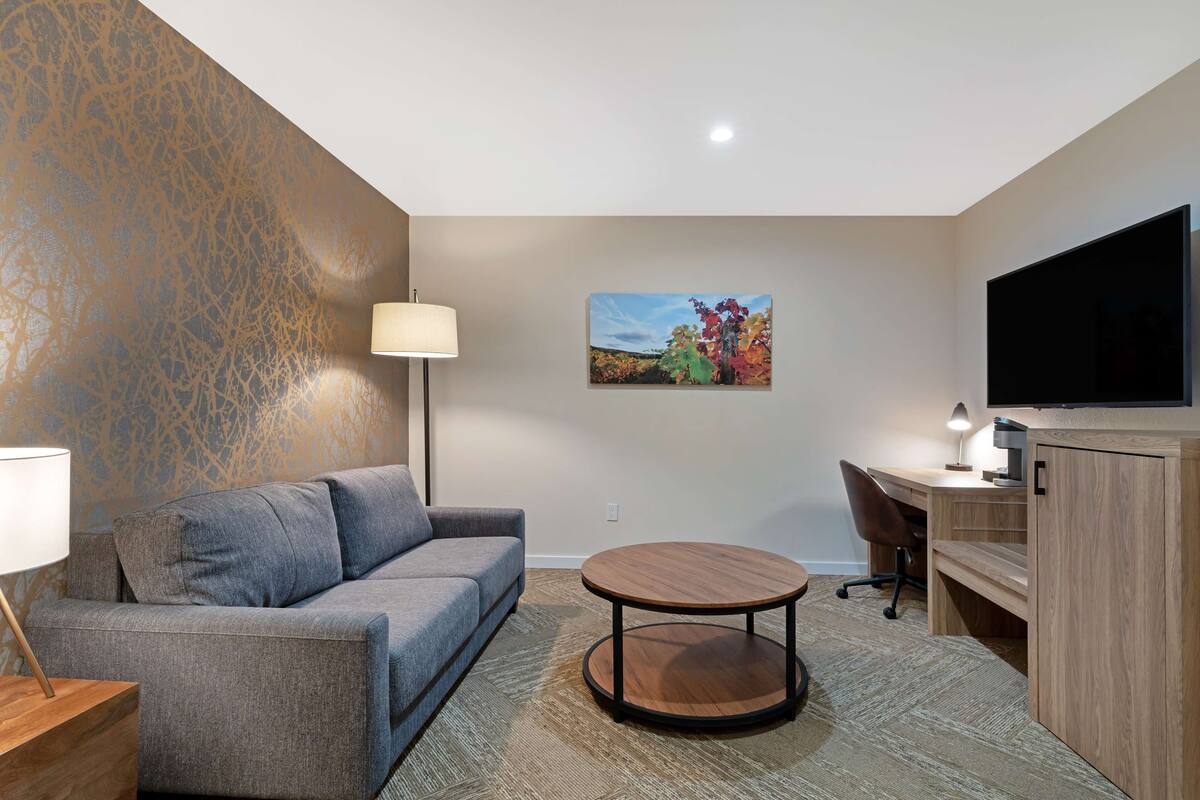 A modern hotel room with a grey sofa, a round wooden coffee table, a desk with a computer, a flat-screen TV, and a painting of autumn leaves on the wall.