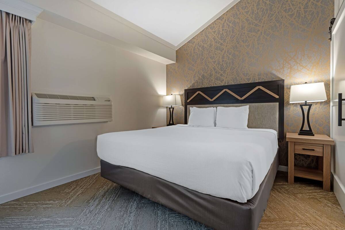 A hotel room with a large bed, two bedside tables with lamps, a wall-mounted air conditioner, and a patterned wallpaper.