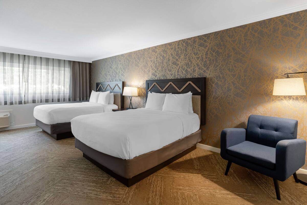 A hotel room with two beds, a blue armchair, and a patterned wall.